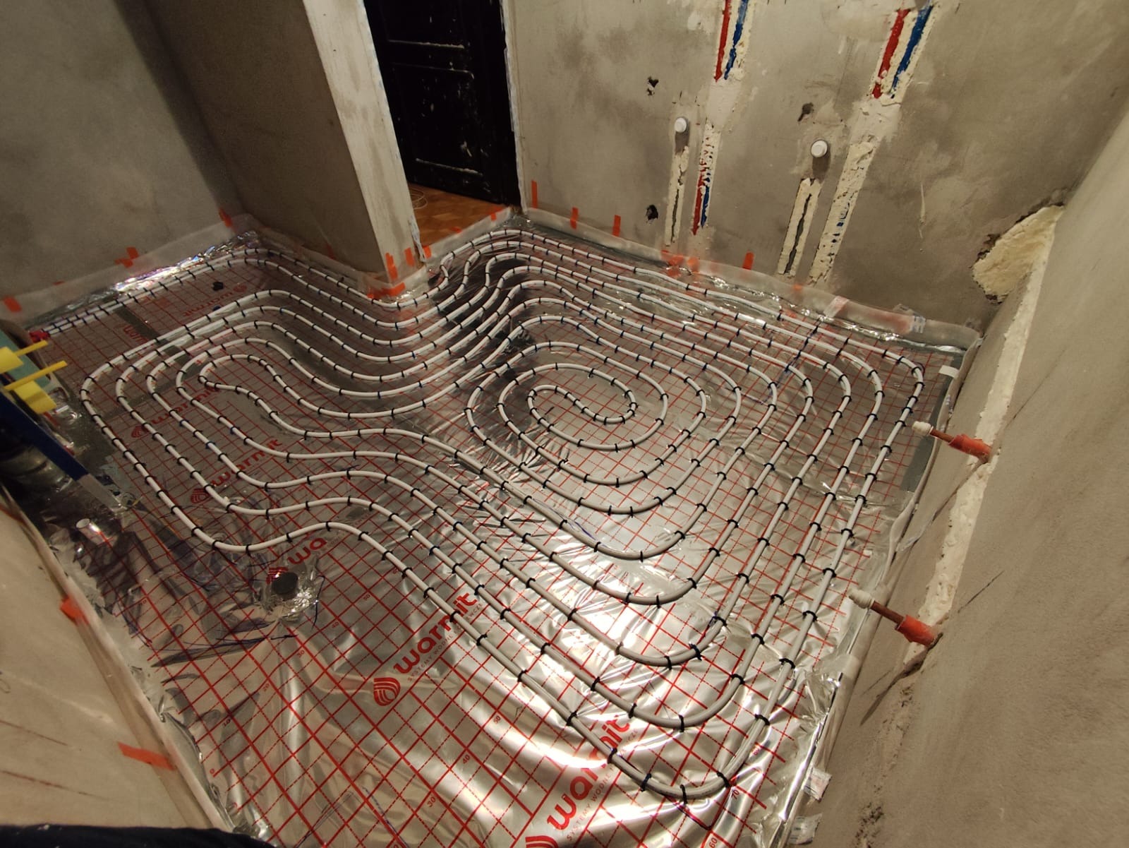 Białe rury ogrzewania podłogowego ułożone spiralnie na folii izolacyjnej z czerwoną siatką w surowym pomieszczeniu łazienki przed wylaniem posadzki.