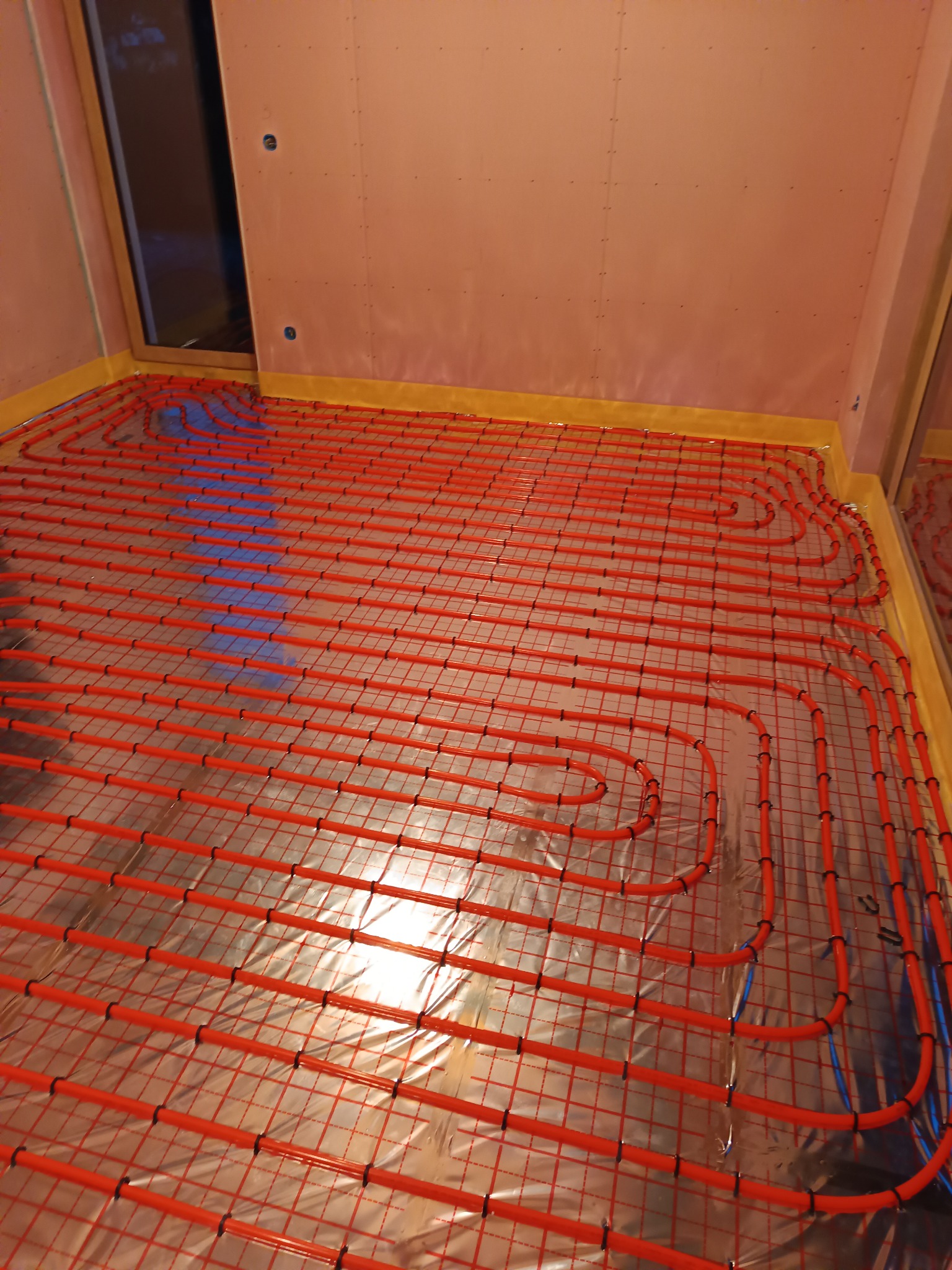 Czerwone rurki ogrzewania podłogowego ułożone na srebrnej folii izolacyjnej w pomieszczeniu z przygotowanymi ścianami.