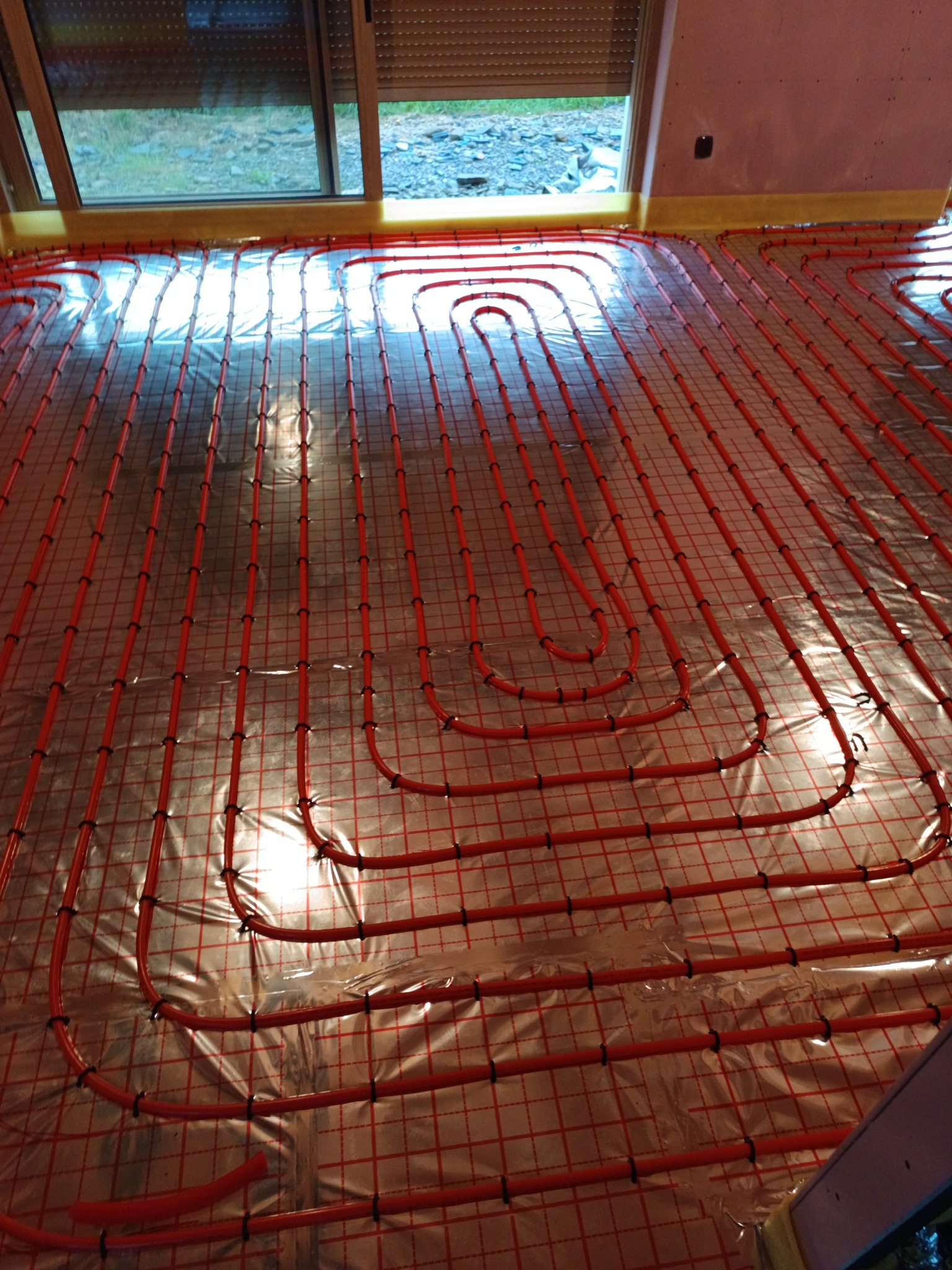 Czerwone rurki ogrzewania podłogowego ułożone na folii z siatką, widoczne okno i fragment ściany