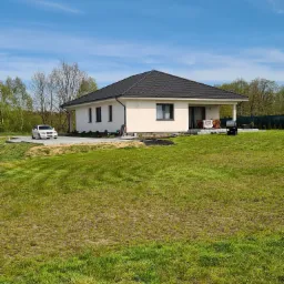 Nowoczesny dom jednorodzinny z ciemną dachówką na tle błękitnego nieba i zielonego trawnika, z białym samochodem zaparkowanym obok.