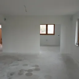 Surowe wnętrze po malowaniu na biało, widoczne ślady na podłodze, okna z drewnianymi ramami i wisząca żarówka na kablu.