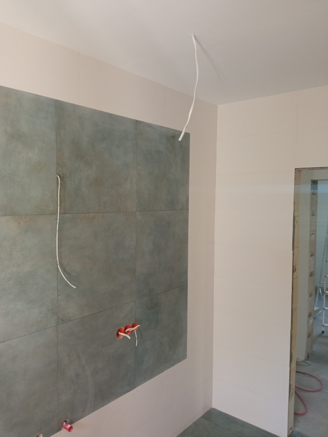 Ściana z szarych, kwadratowych płytek w trakcie montażu, widoczne kable elektryczne wystające ze ściany i sufitu, oraz niezamaskowane puszki elektryczne.