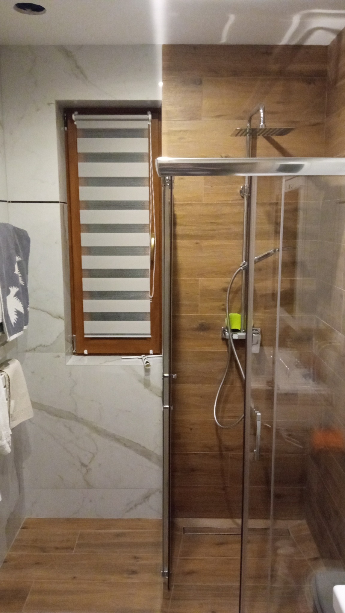 Nowoczesna łazienka z marmurowymi i drewnopodobnymi płytkami, kabiną prysznicową typu walk-in i roletą okienną w brązowej ramie.