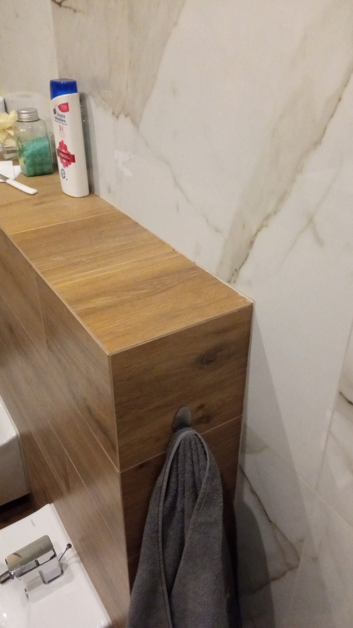 Wykończona łazienka w Berejowie z drewnopodobną zabudową umywalki i marmurowymi płytkami na ścianie. Szary ręcznik zawieszony na haczyku.