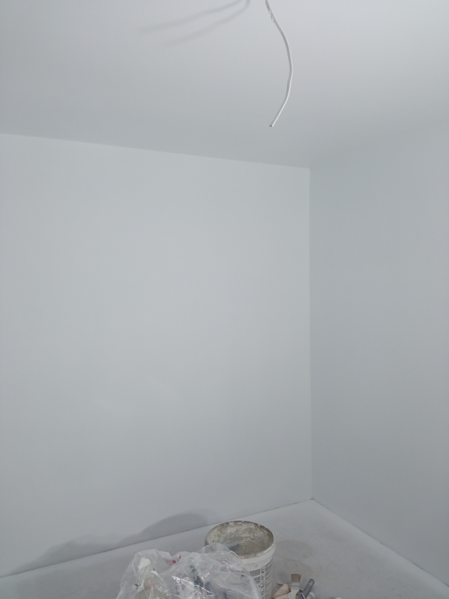 Świeżo pomalowane na biało wnętrze pokoju, widoczny wystający kabel z sufitu i wiadro z farbą na podłodze, etap prac wykończeniowych.