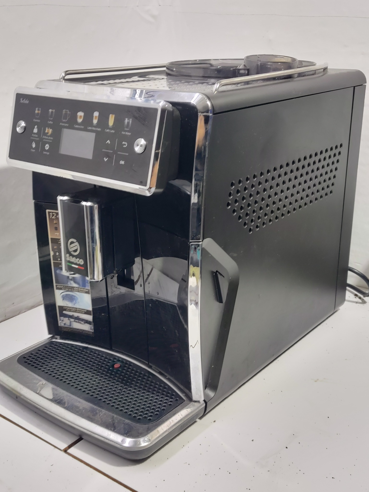 Automatyczny ekspres do kawy Saeco w czarnej obudowie z chromowanymi detalami, widoczny panel sterowania z ikonami różnych napojów, na białym tle.