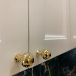 Białe fronty szafek z frezowanymi krawędziami i okrągłymi, złotymi gałkami na tle blatu z czarnego marmuru.