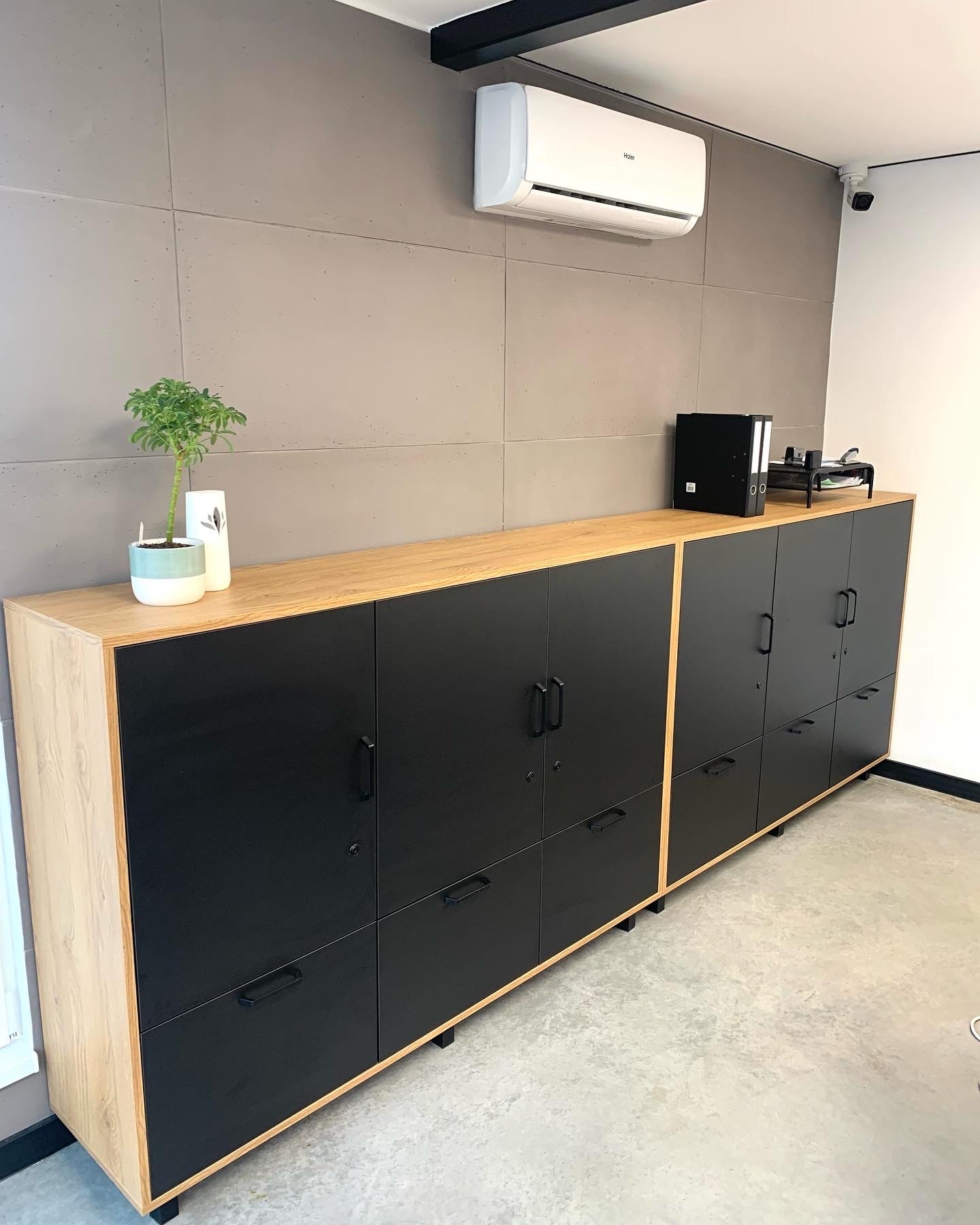 Długa szafa biurowa na wymiar z czarnymi frontami i blatem z jasnego drewna, na której stoi roślina doniczkowa i czarny segregator, na tle szarej ściany z klimatyzatorem.