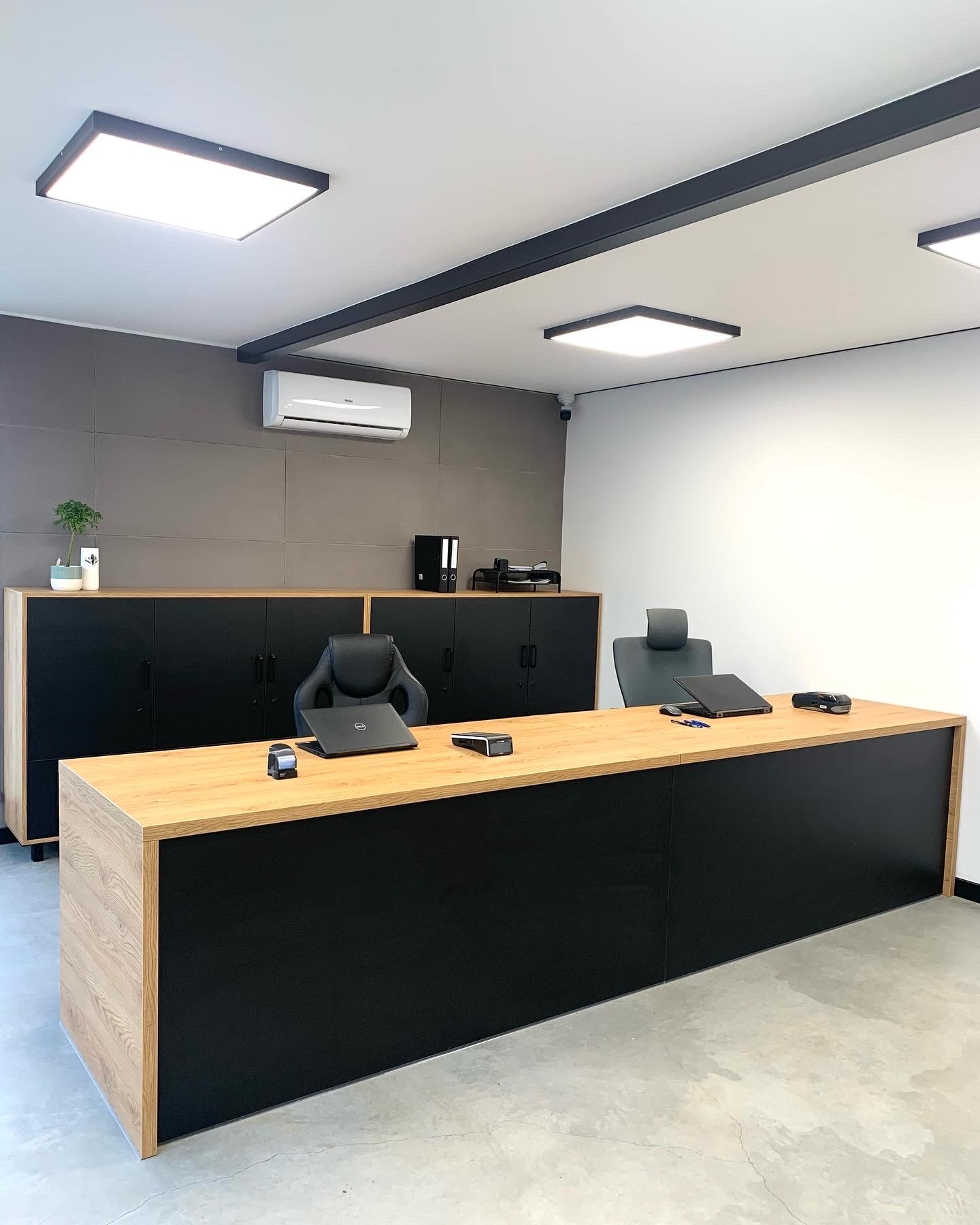Nowoczesne biuro z dużym biurkiem z blatem drewnianym i czarnym frontem, czarne szafki i szara ściana w tle, minimalistyczny wystrój.