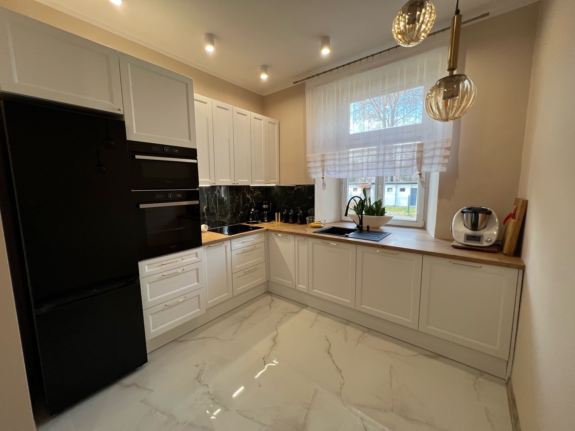 Nowoczesna kuchnia w bieli z drewnianym blatem, czarnym AGD i marmurowym backsplashu, oświetlona wiszącymi lampami i światłem dziennym z okna.