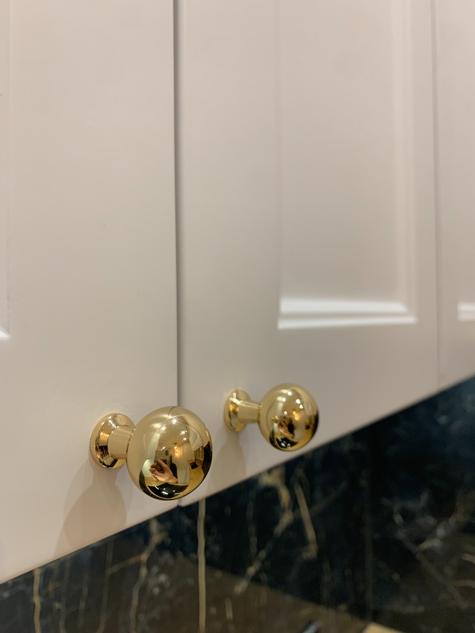 Białe fronty szafek z frezowanymi krawędziami i okrągłymi, złotymi gałkami na tle blatu z czarnego marmuru.