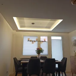 Nowoczesna jadalnia z podwieszanym sufitem z oświetleniem LED w ramie, szarymi krzesłami i drewnianym stołem, nad którym wisi designerska lampa z kulistymi kloszami. Na stole bukiet kwiatów...