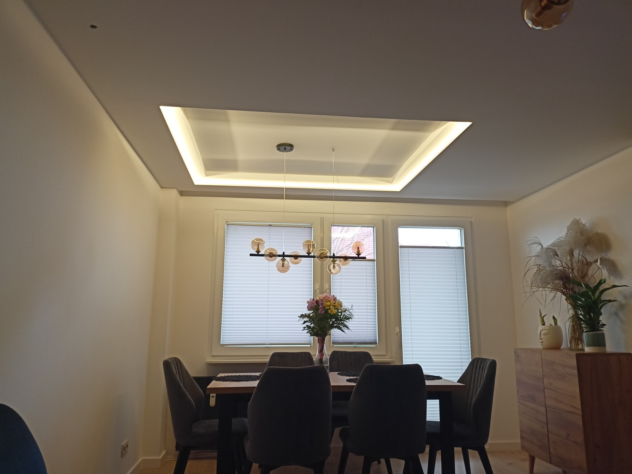 Nowoczesna jadalnia z podwieszanym sufitem z oświetleniem LED w ramie, szarymi krzesłami i drewnianym stołem, nad którym wisi designerska lampa z kulistymi kloszami. Na stole bukiet kwiatów...