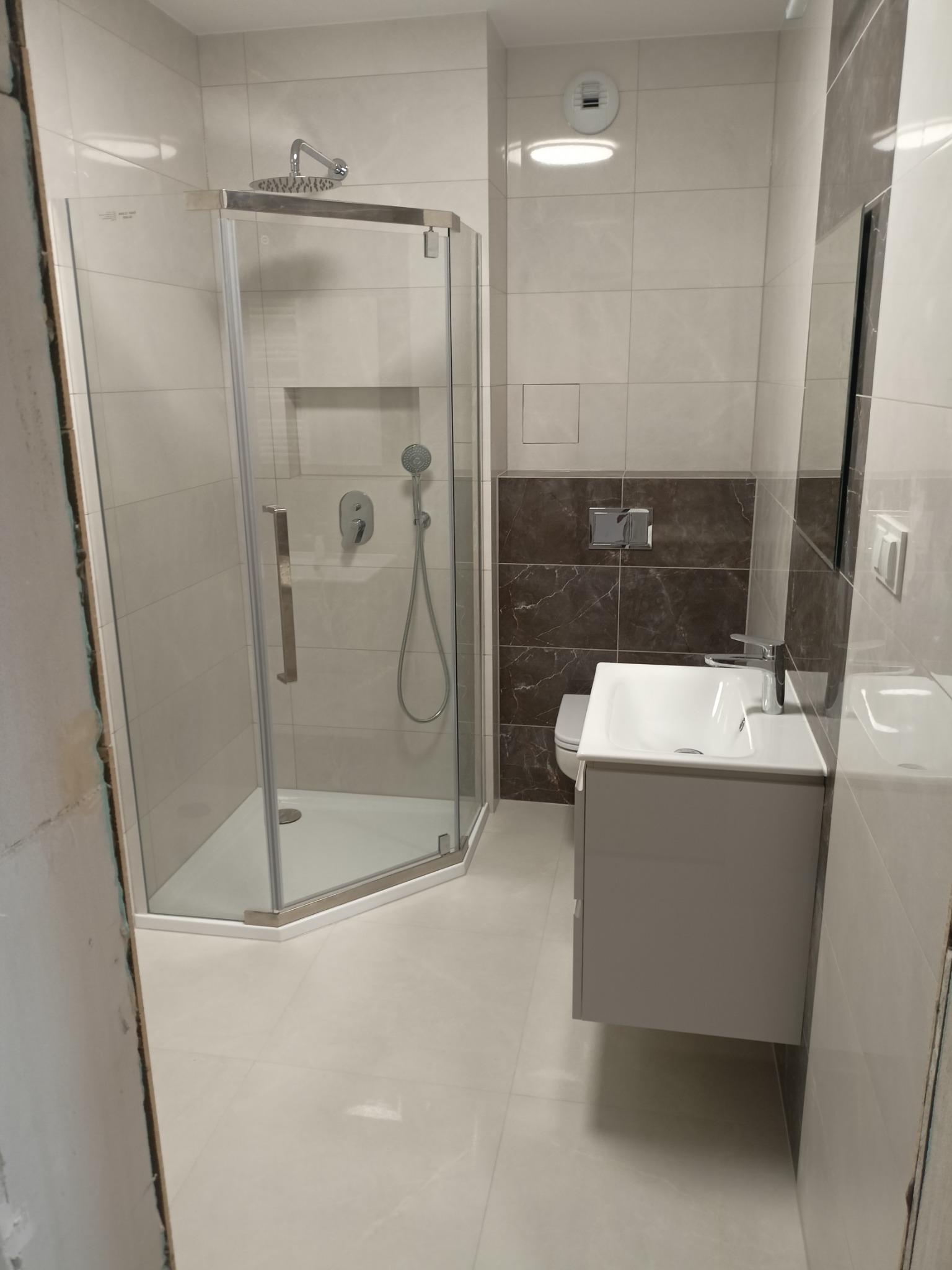 Wykończona, nowoczesna łazienka w jasnych płytkach z kabiną prysznicową typu walk-in, szarą szafką pod umywalką i fragmentem ściany z ciemnobrązowych płytek imitujących marmur.