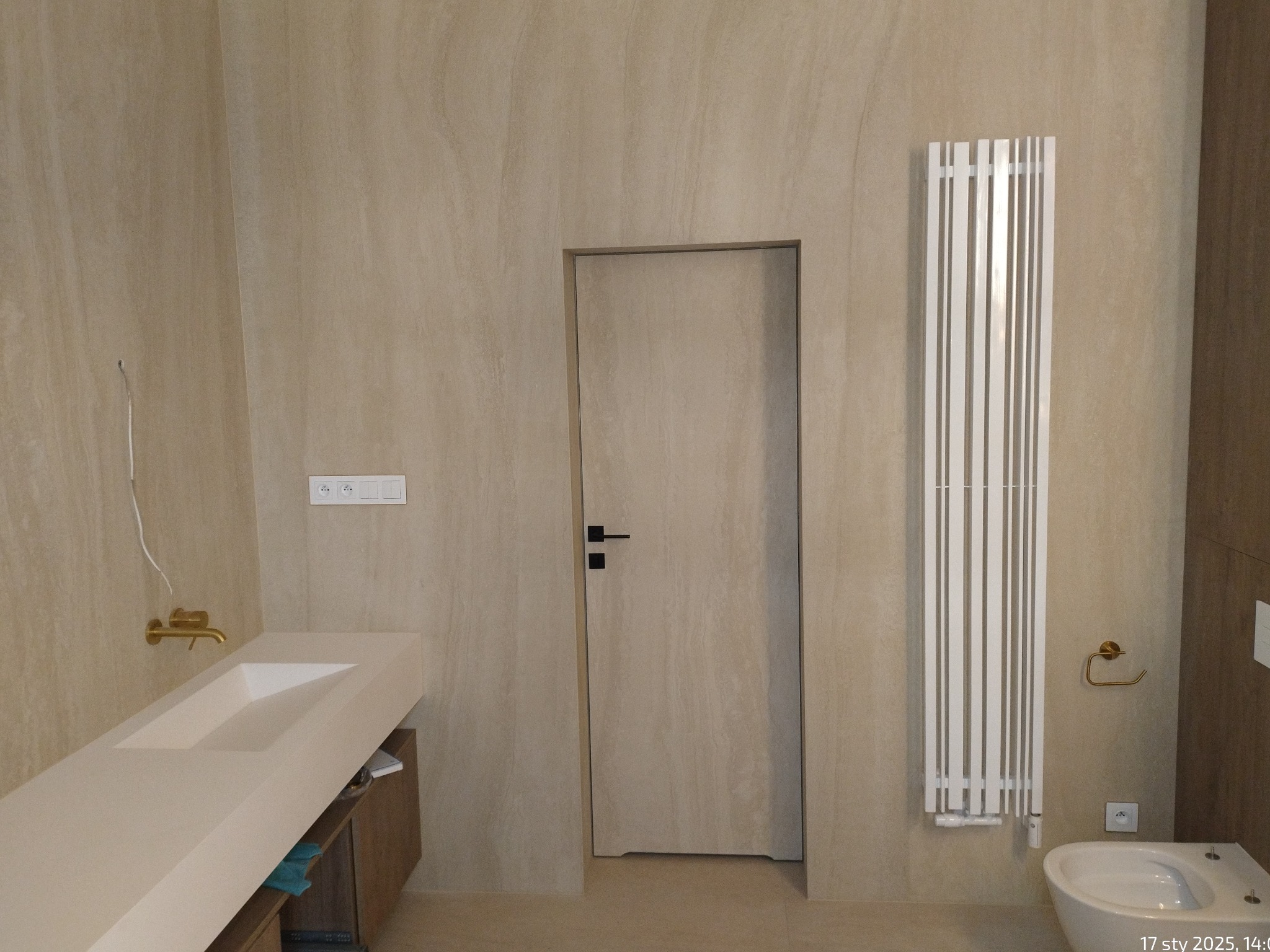 Beżowa łazienka z umywalką, grzejnikiem i toaletą. Minimalistyczny design, neutralna kolorystyka, nowoczesne wykończenie. Widoczne detale armatury i oświetlenia.