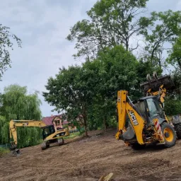 Dwie żółte maszyny budowlane: koparka CAT 319D oraz koparko-ładowarka JCB, na placu budowy z przygotowanym terenem pod budowę, w tle drzewa i dom z czerwoną dachówką.