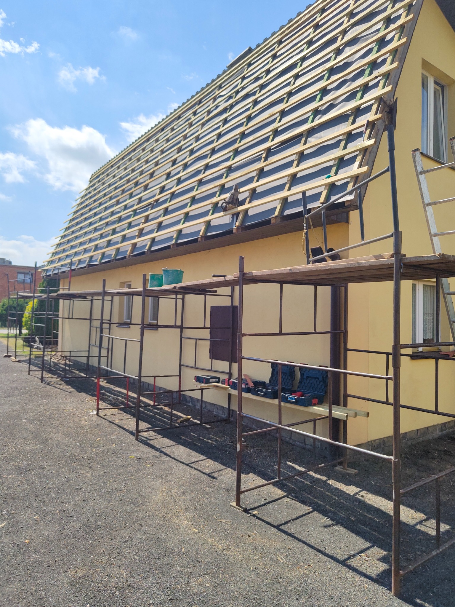Żółty budynek w trakcie prac dekarskich, widoczne rusztowania, narzędzia i przygotowana konstrukcja pod pokrycie dachu.
