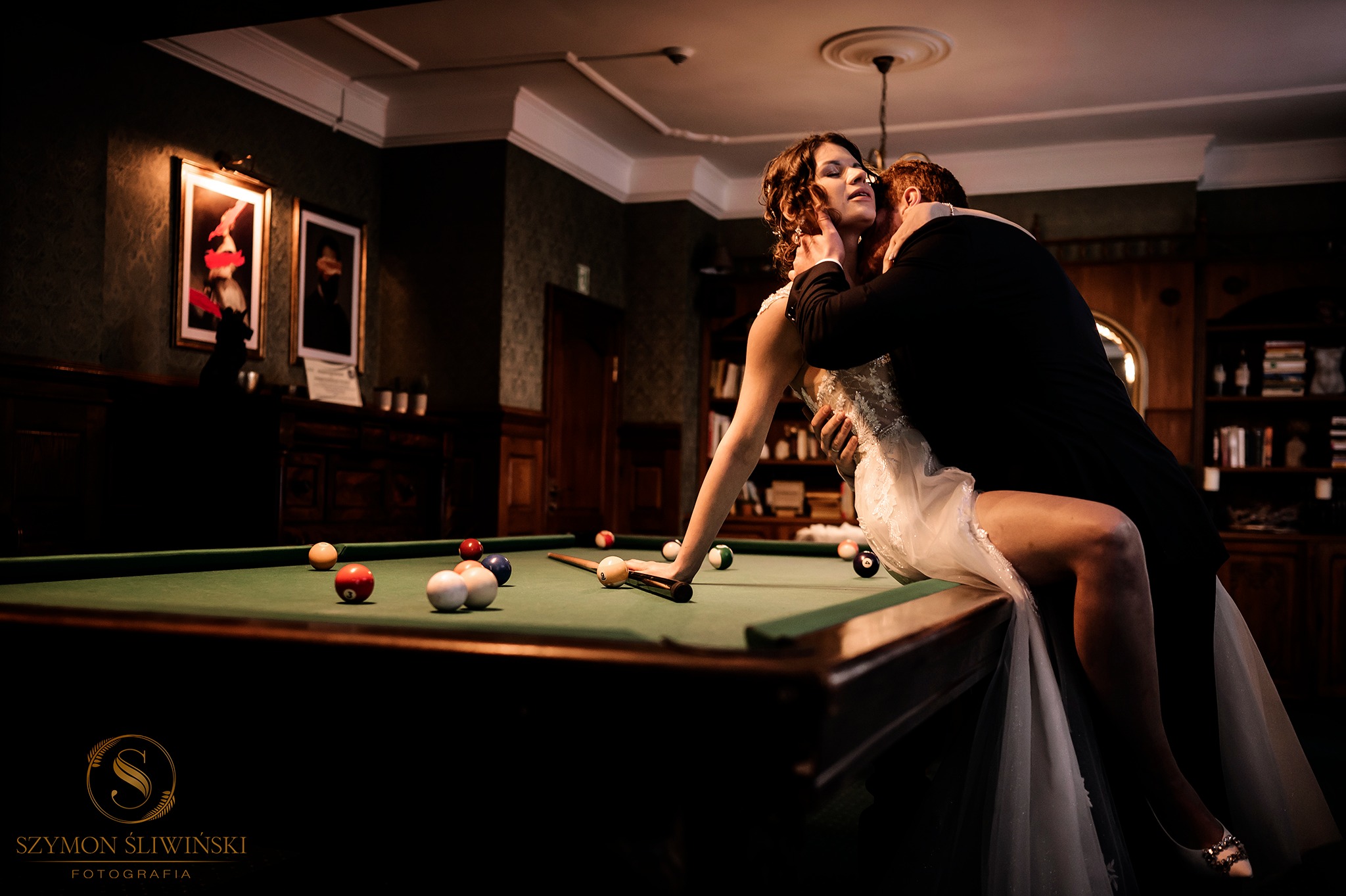 Panna młoda w koronkowej sukni siedzi na stole bilardowym, obejmowana przez pana młodego w eleganckim smokingu w ciemnym pomieszczeniu z biblioteczką w tle.