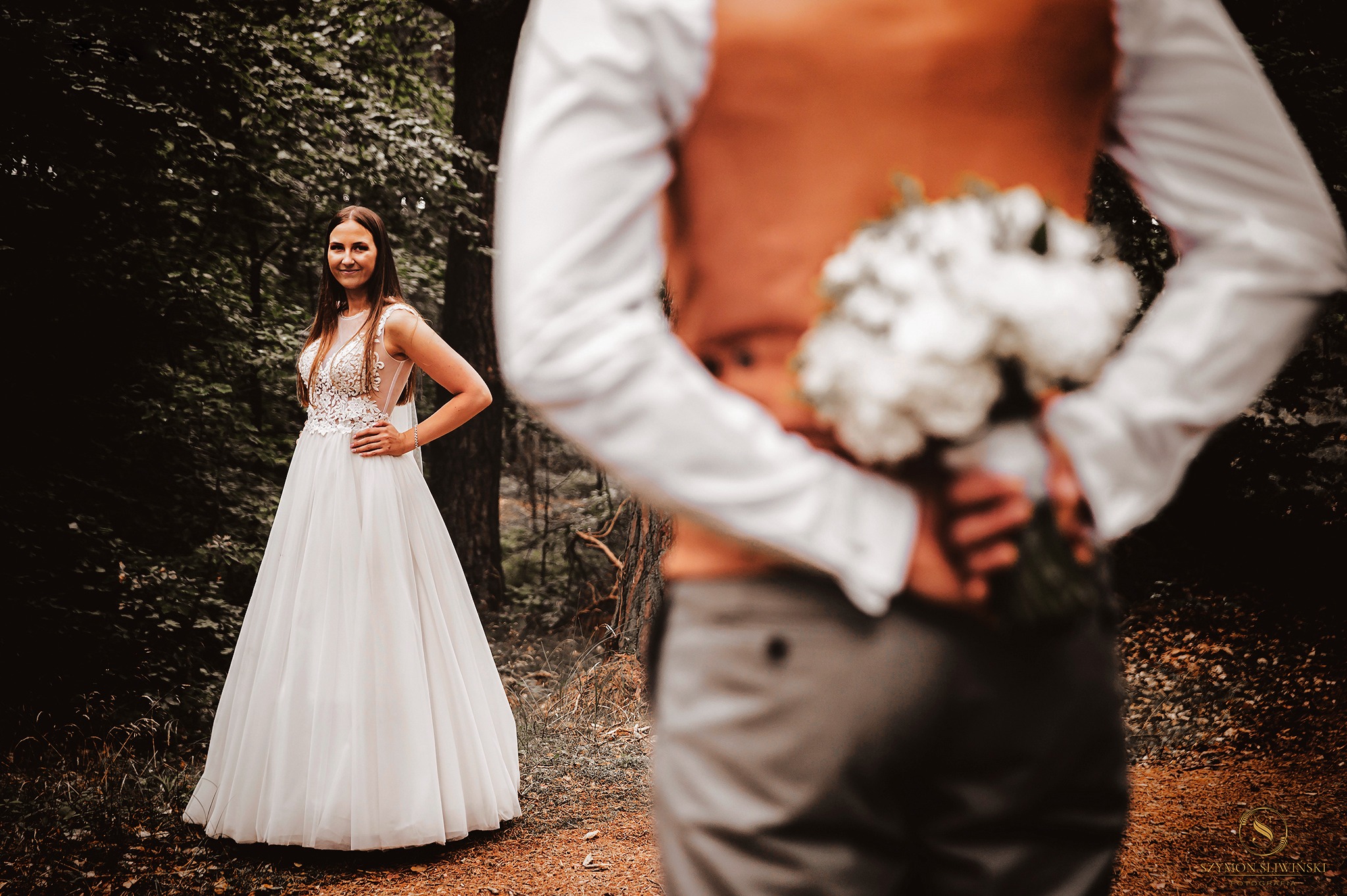 Panna młoda w białej sukni z koronkową górą, pozująca w lesie, podczas gdy pan młody trzyma za plecami bukiet białych kwiatów.