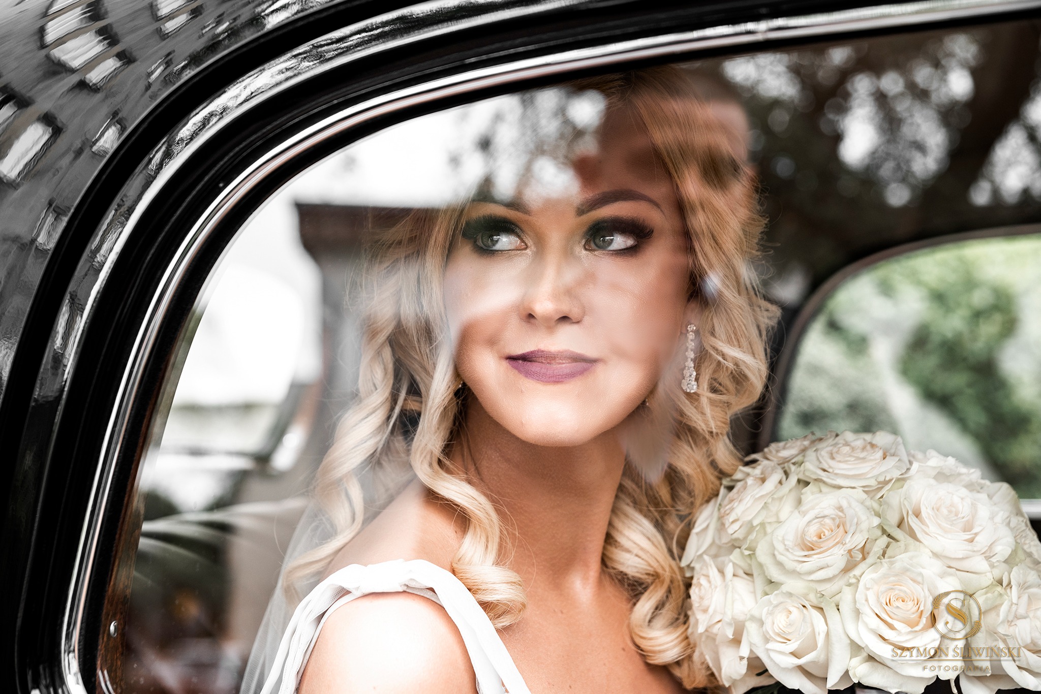 Panna młoda z blond lokami i bukietem białych róż spogląda przez okno czarnego, klasycznego samochodu.