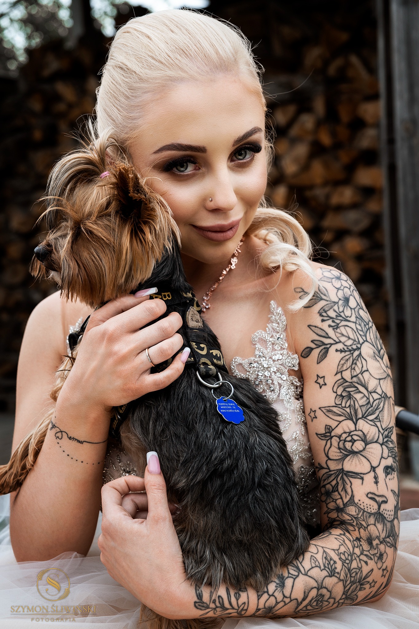 Uśmiechnięta blondynka w sukni zdobionej kryształkami, trzymająca na rękach yorka z niebieską adresówką, widoczne tatuaże na ramieniu i drewno w tle.