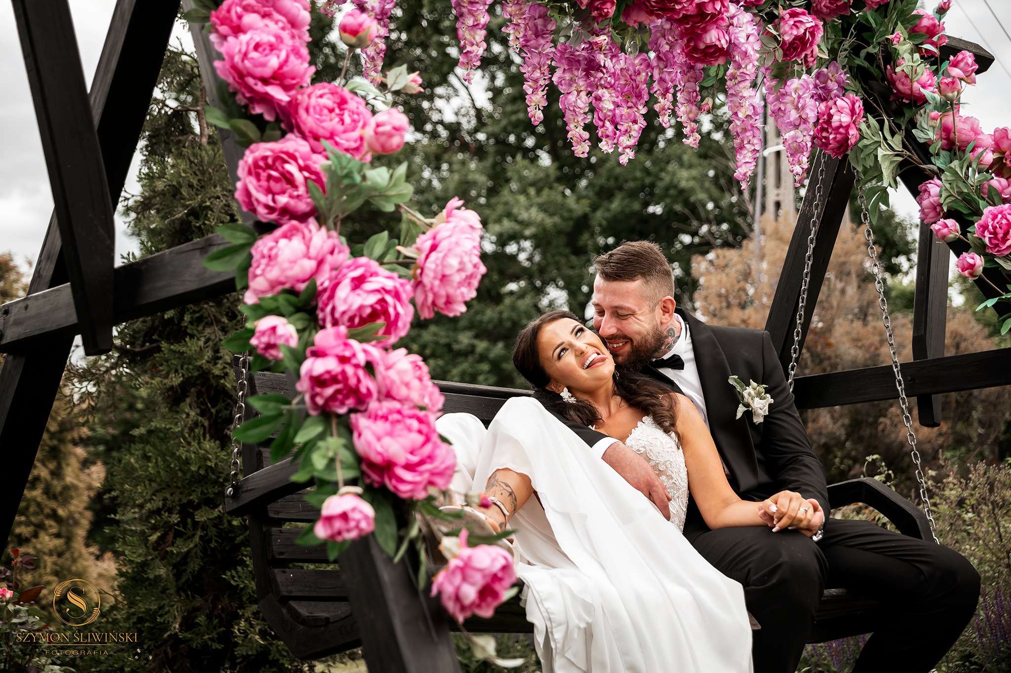 Uśmiechnięta para młoda w strojach ślubnych siedzi na czarnej huśtawce ozdobionej różowymi i fioletowymi kwiatami w plenerze.