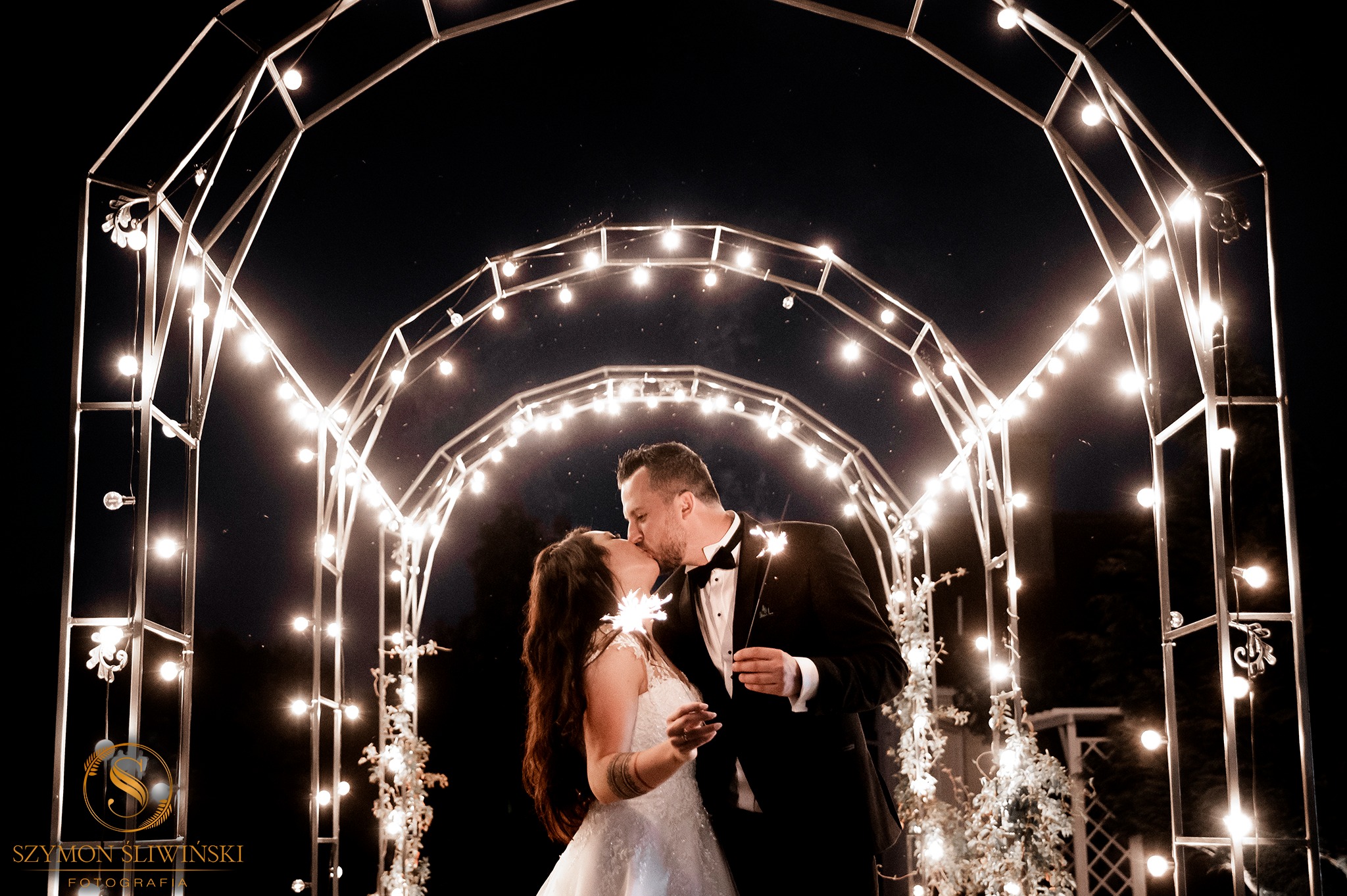 Pocałunek pary młodej w nocy, trzymającej zimne ognie, pod metalową konstrukcją z żarówkami, panna młoda z tatuażem na ramieniu.