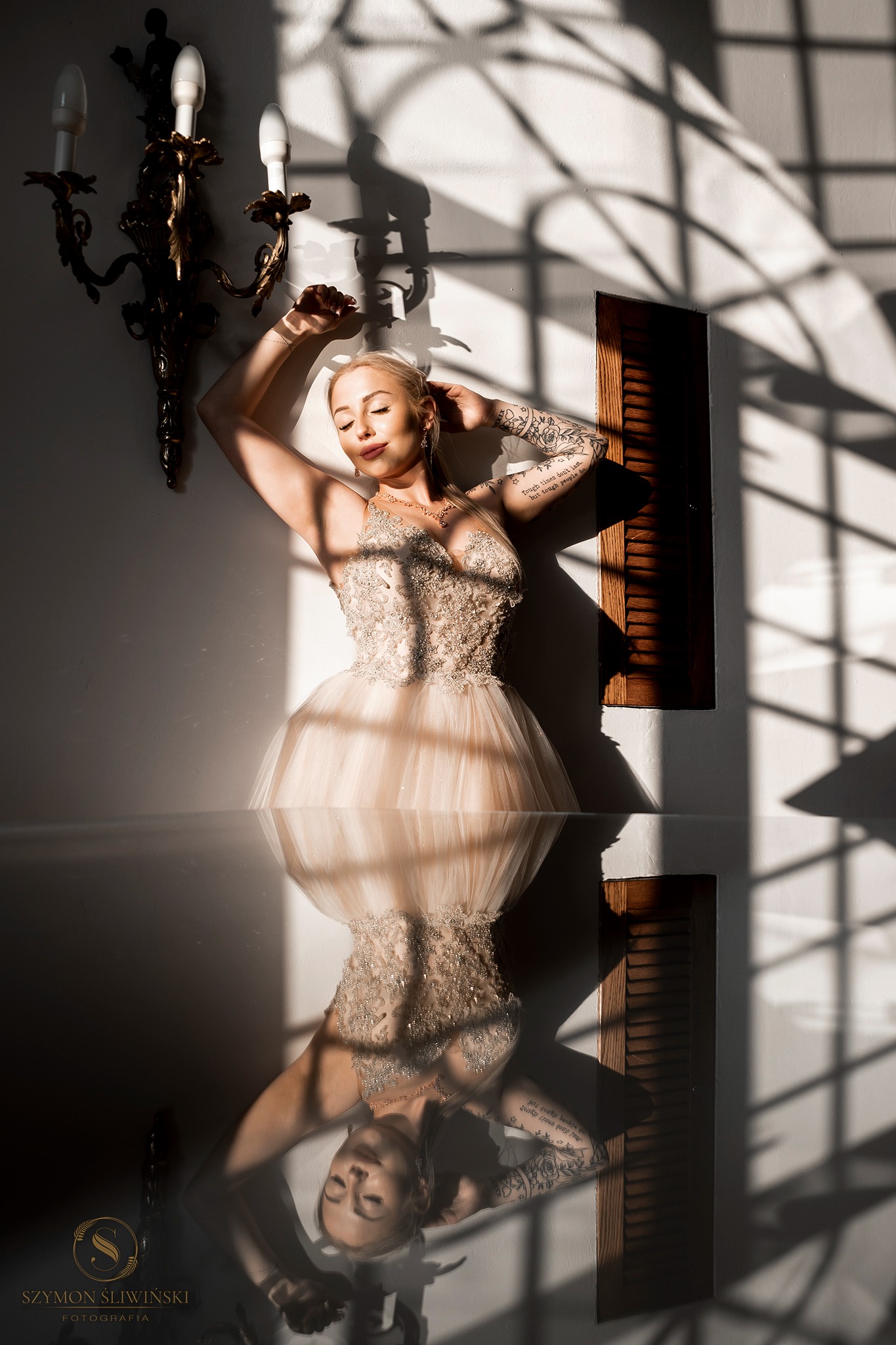 Kobieta w balowej sukni pozuje pod ścianą w plamach słonecznego światła, widoczne tatuaże na ramieniu i odbicie w szklanej powierzchni poniżej.