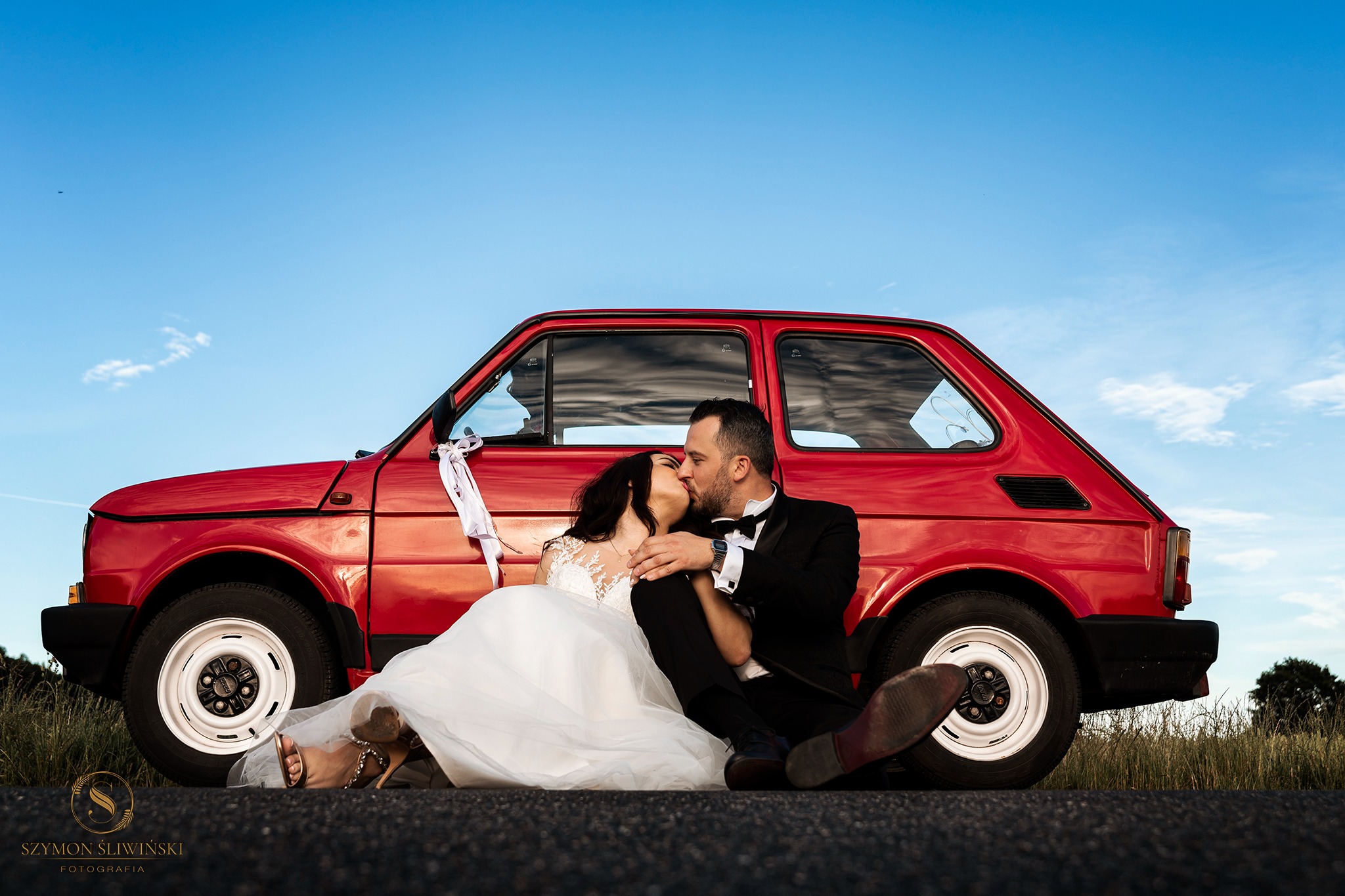 Pocałunek młodej pary siedzącej przed czerwonym Fiatem 126p z białymi wstążkami na klamce, na tle błękitnego nieba.