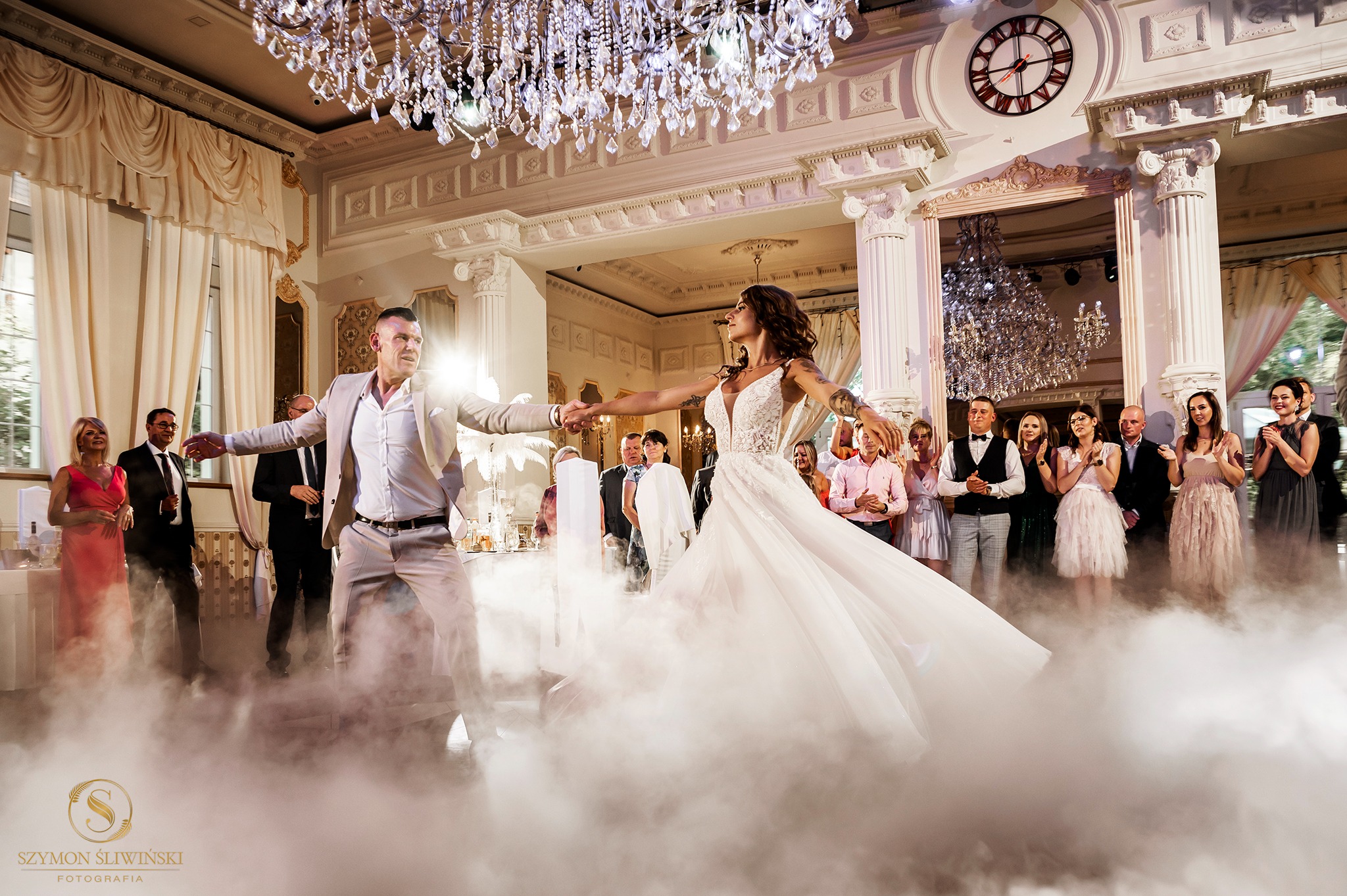 Pierwszy taniec pary młodej w eleganckiej sali balowej z kryształowym żyrandolem, oświetlony sztuczną mgłą i oklaskiwany przez gości.