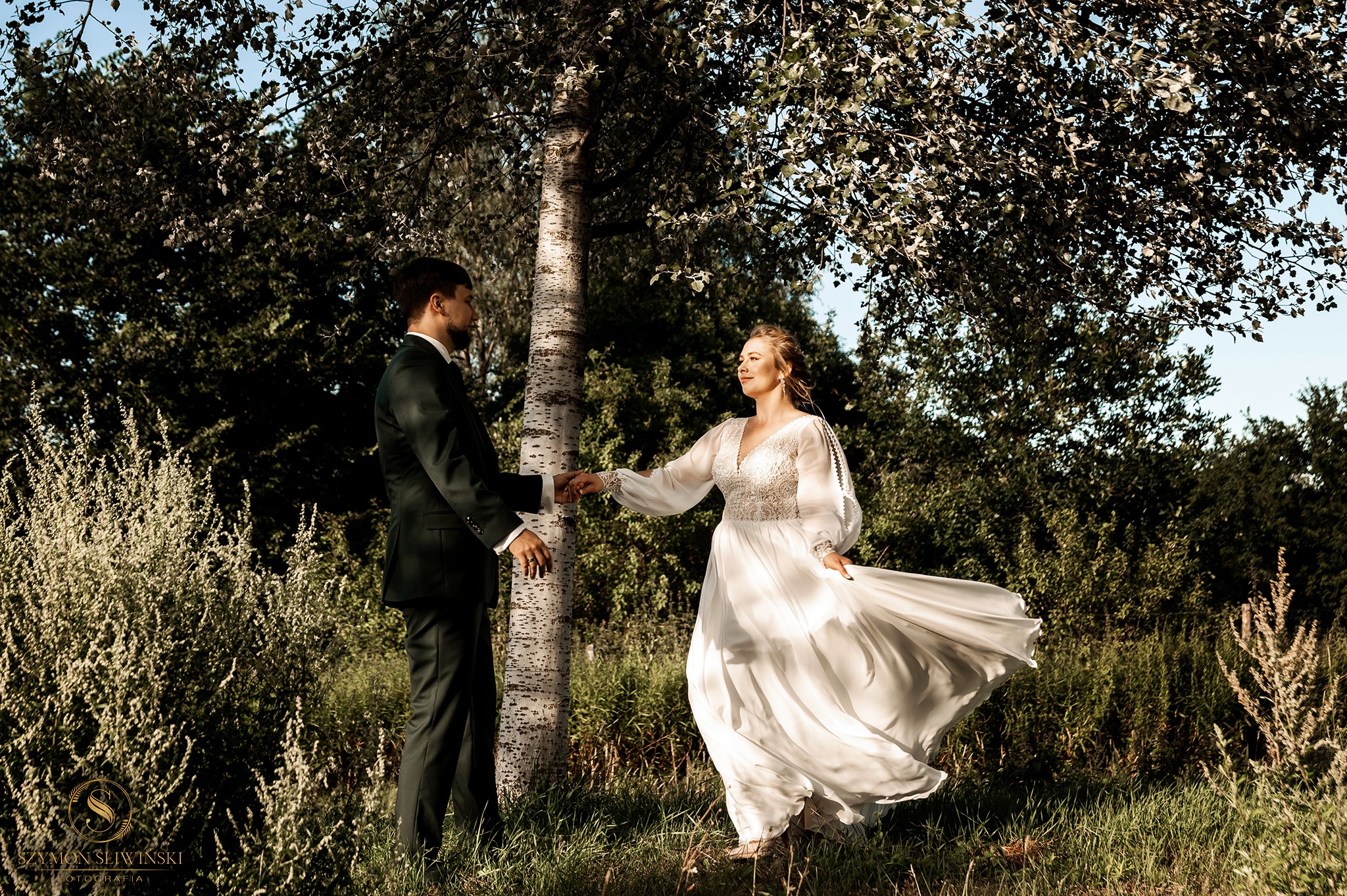 Para młoda trzymająca się za ręce przy brzozie na tle zieleni. Suknia panny młodej powiewa na wietrze.