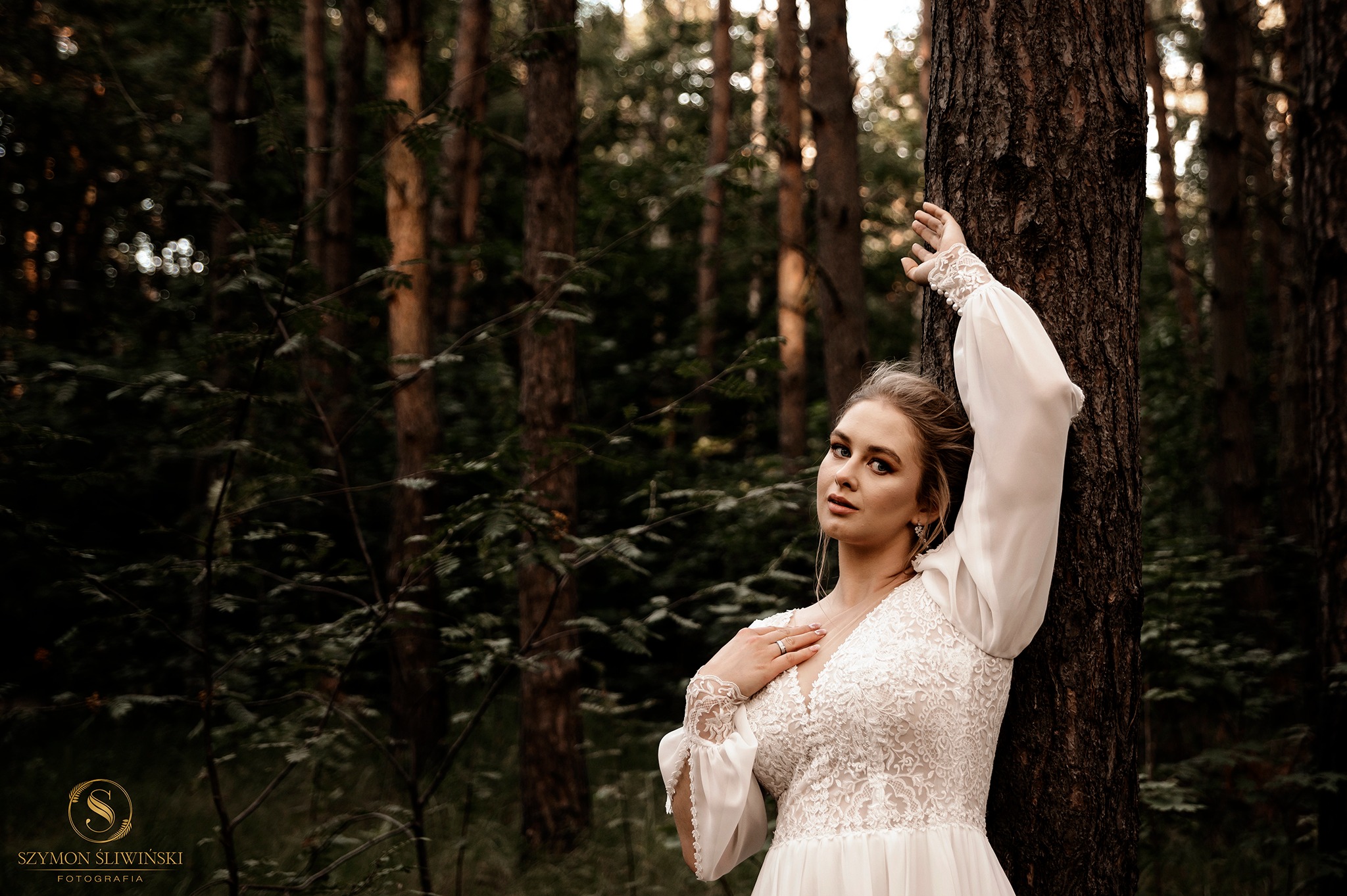 Portret kobiety w białej sukni z koronkową górą, pozującej przy drzewie w lesie, z delikatnym makijażem i upiętymi włosami.