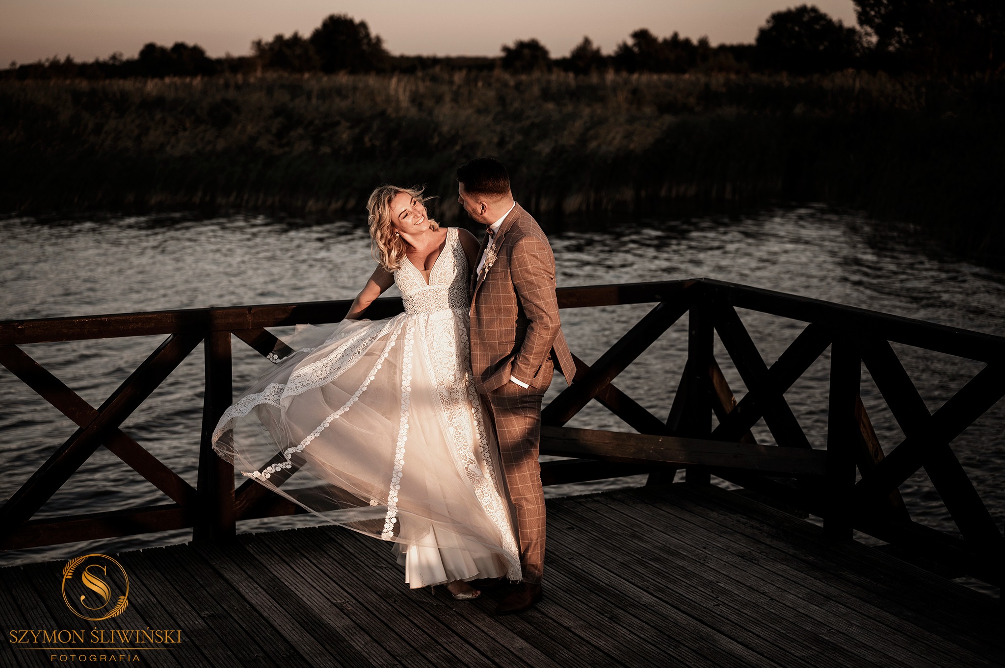 Para młoda na drewnianym pomoście nad jeziorem o zachodzie słońca. Panna młoda w koronkowej sukni z tiulowym dołem, odwraca się z uśmiechem do pana młodego w brązowym garniturze w kratę.