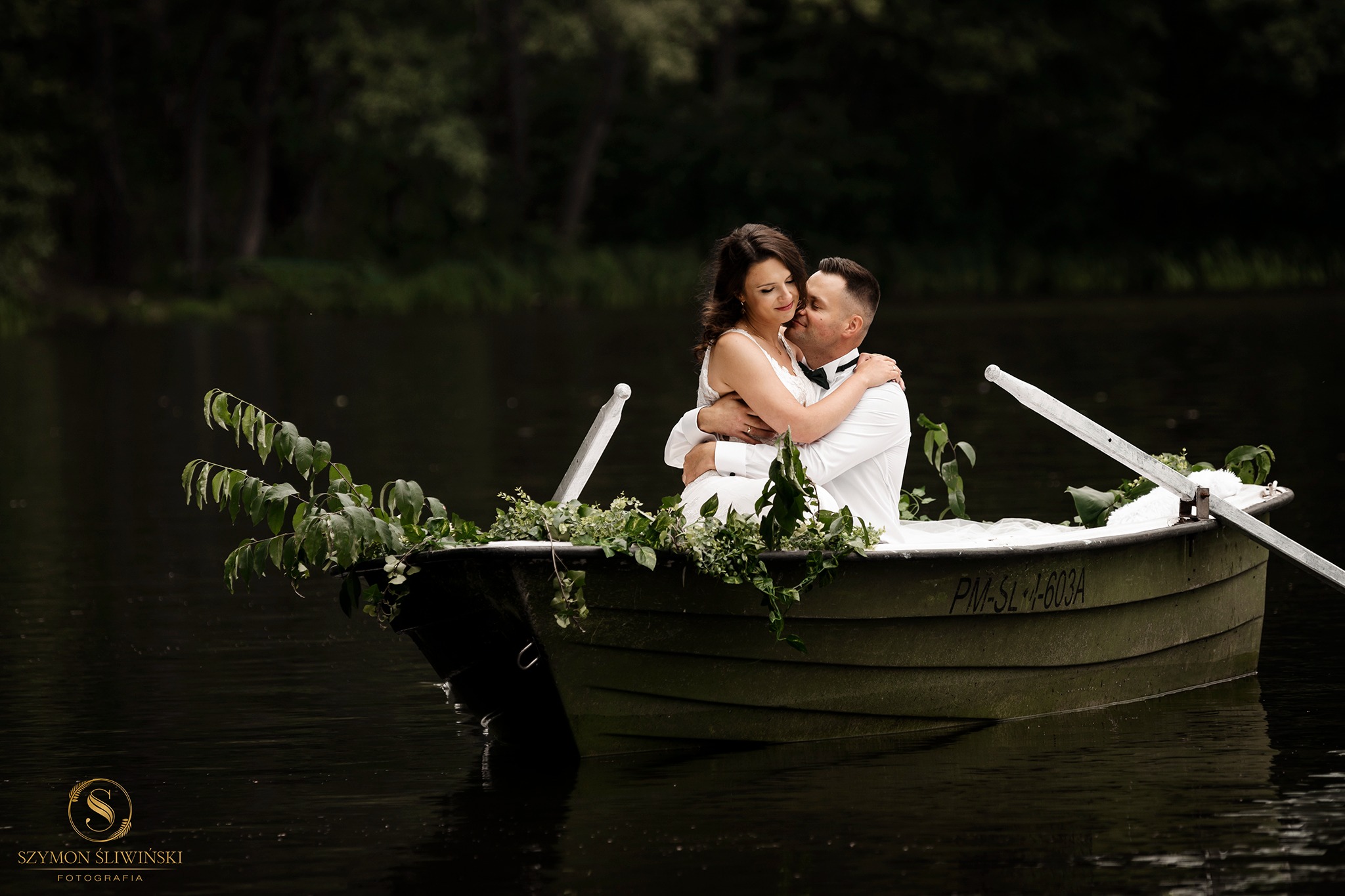 Para młoda w objęciach siedzi w łódce na jeziorze, łódka ozdobiona zielonymi gałązkami, ciemne tło drzew.