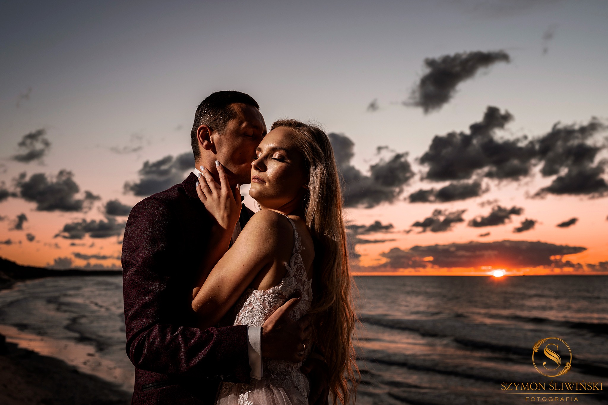 Para młoda w objęciach na tle zachodzącego słońca nad morzem, panna młoda w koronkowej sukni, pan młody w bordowym garniturze z wzorem.