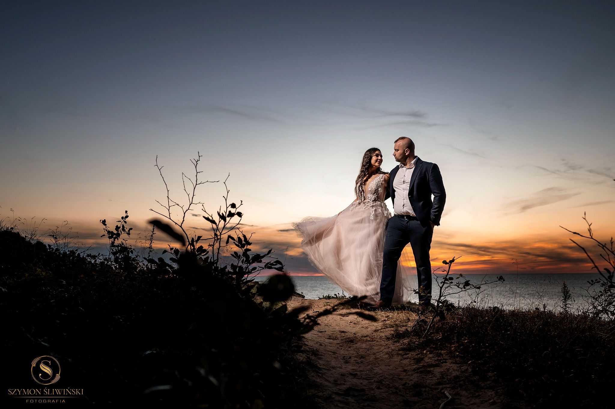 Para młoda w sukni ślubnej i garniturze stoi na wzgórzu z widokiem na zachodzące słońce nad morzem, z siluetami roślin na pierwszym planie.