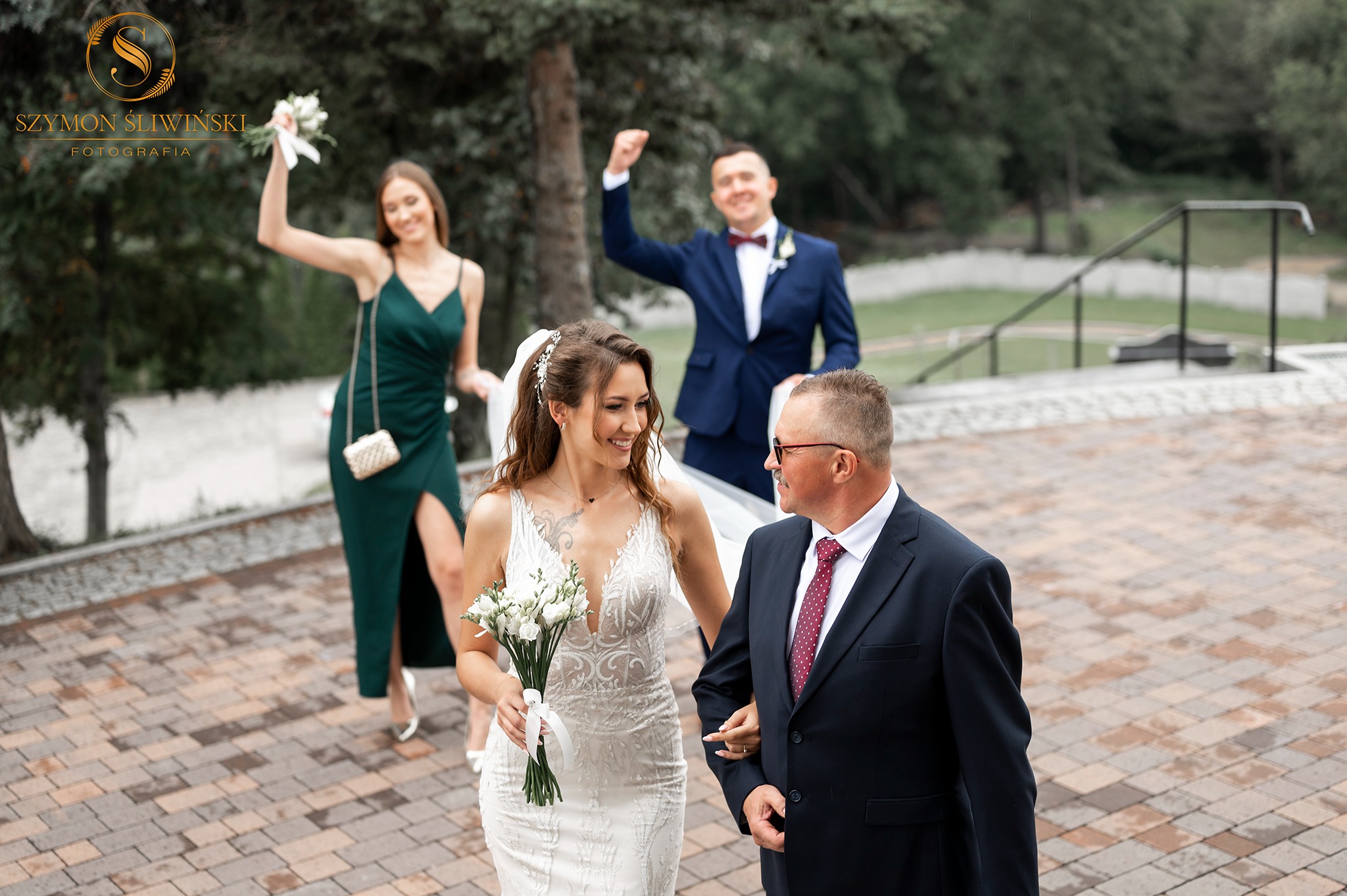 Uśmiechnięta panna młoda w koronkowej sukni prowadzona przez ojca, w tle drużba z podniesioną ręką i druhna w zielonej sukience trzymająca bukiet