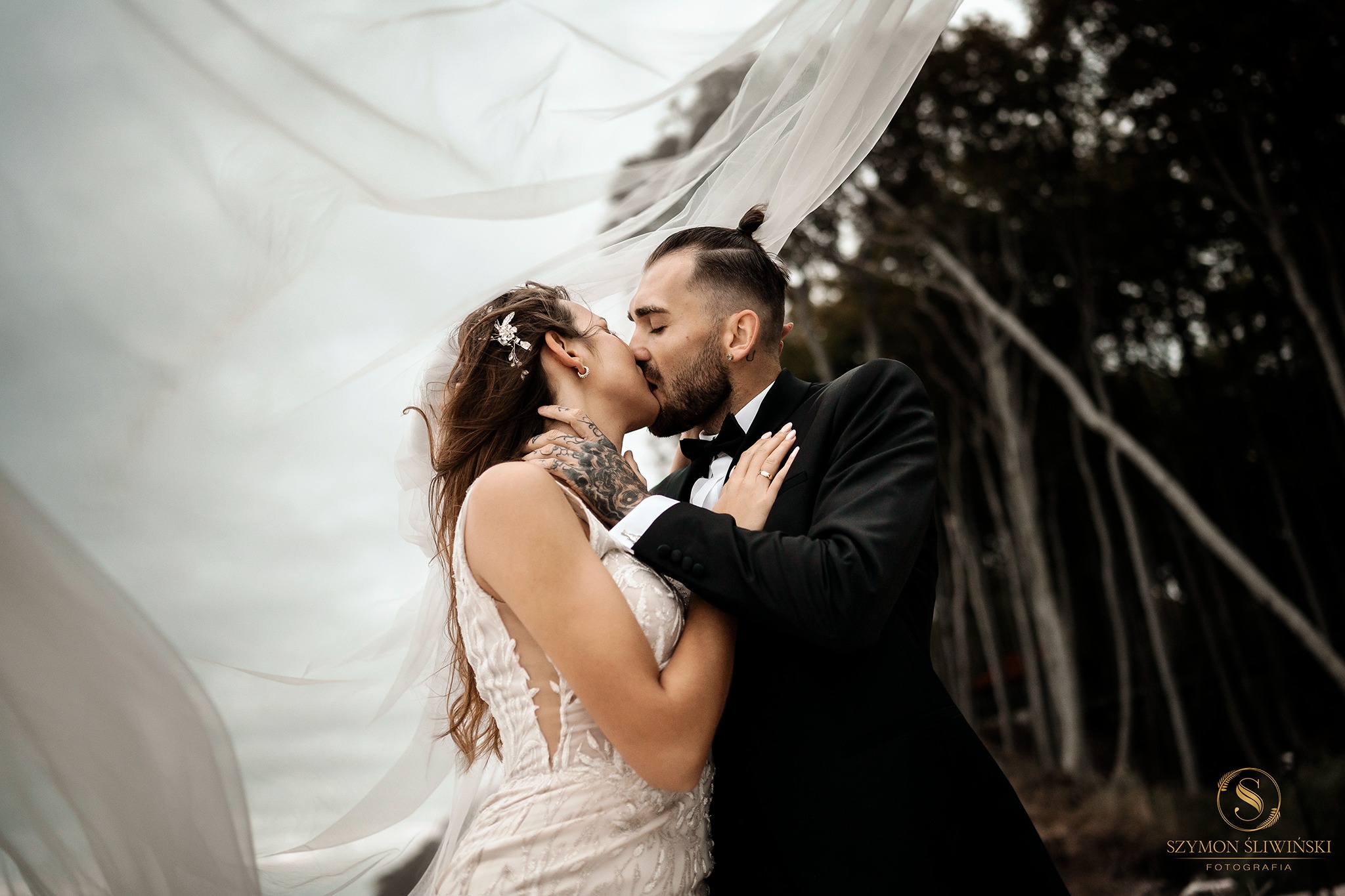 Para młoda całuje się pod rozwianym welonem na tle lasu, panna młoda w sukni z koronkowymi detalami i ozdobą we włosach, pan młody w czarnym smokingu z muszką, widoczne tatuaże na dłoni panny młodej.