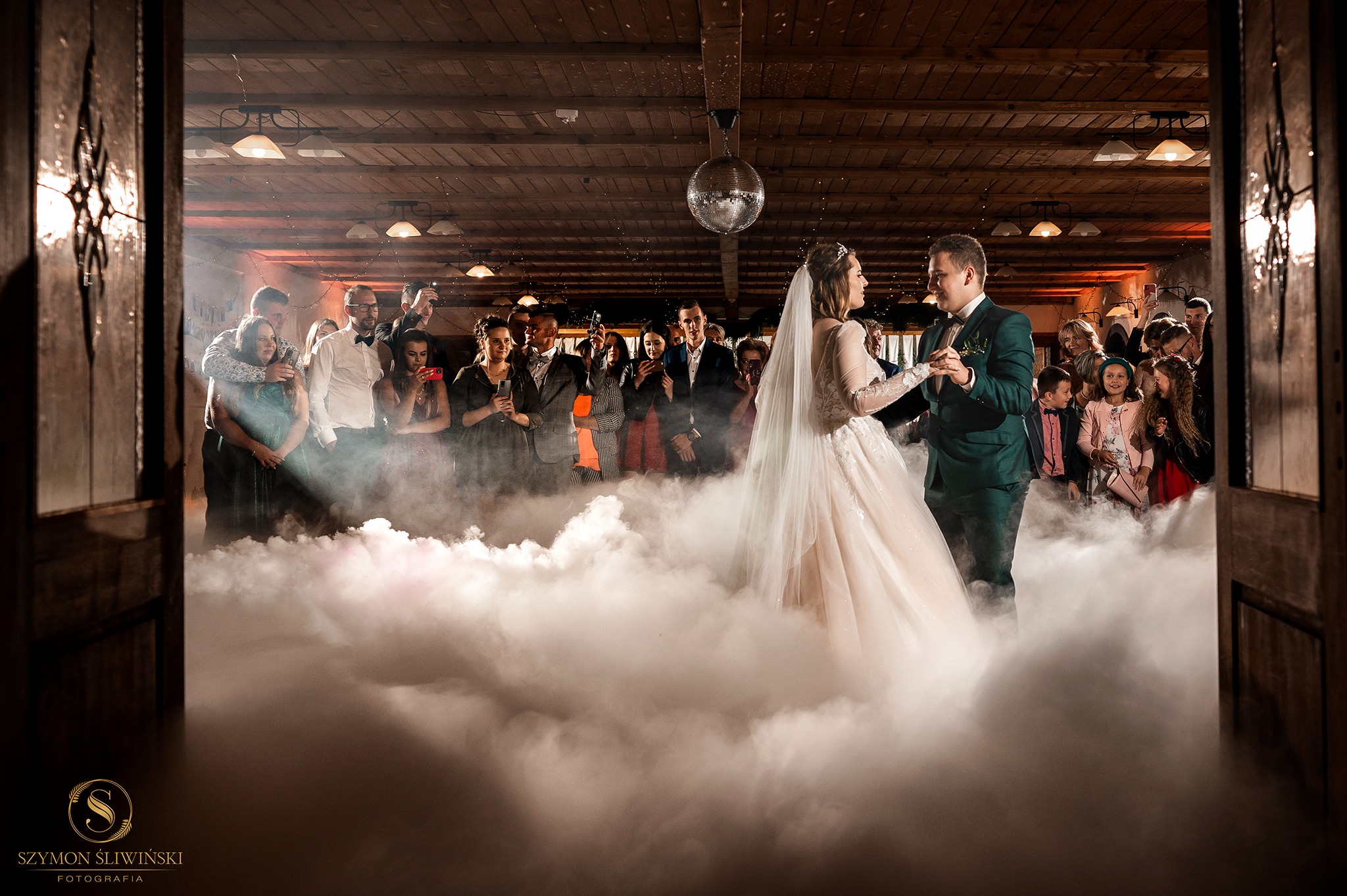 Para młoda tańczy pierwszy taniec w dymie na sali weselnej z drewnianym stropem i lustrzaną kulą, goście obserwują zza otwartych drzwi.