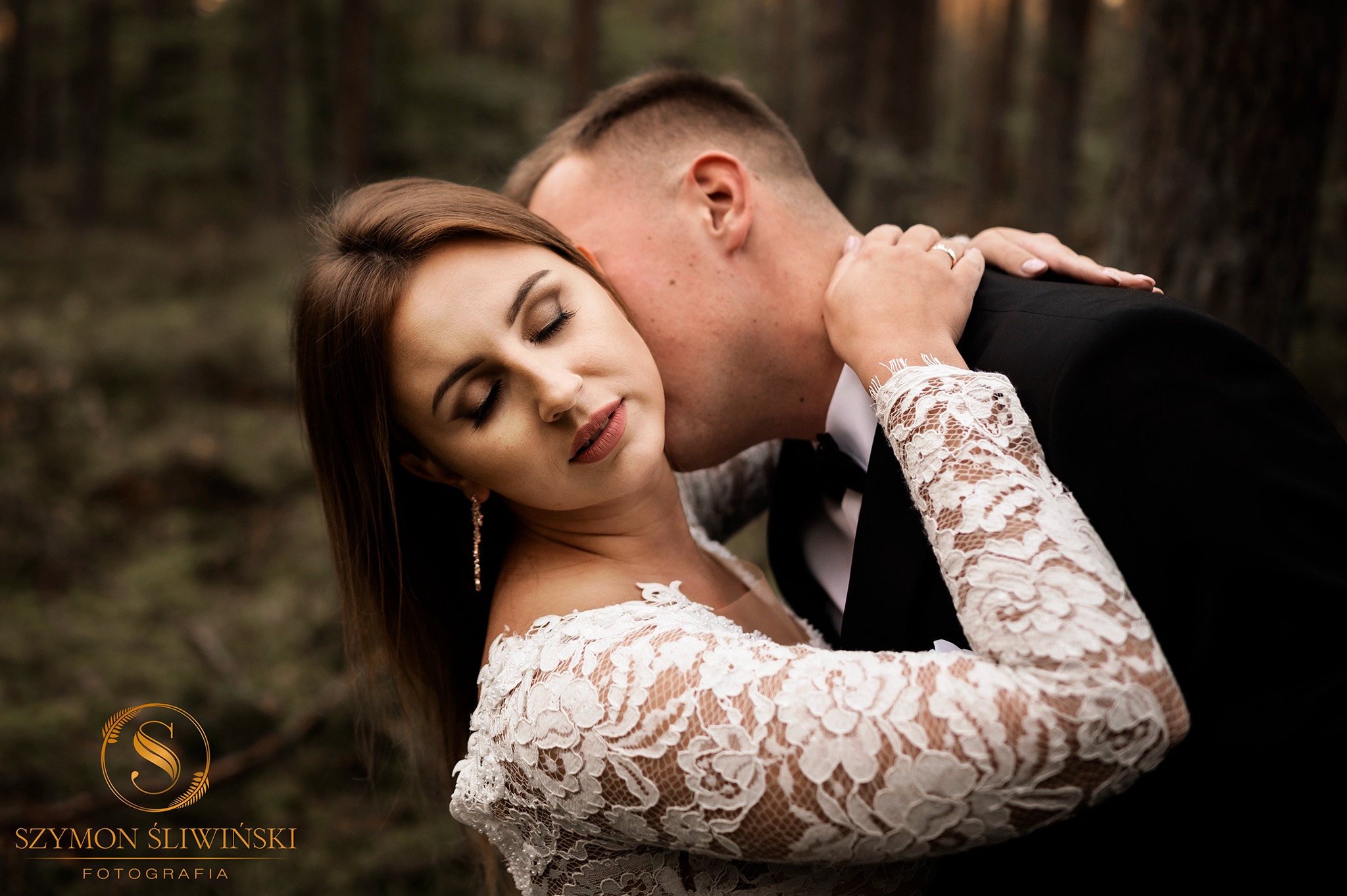 Panna młoda w koronkowej sukni z długim rękawem, z zamkniętymi oczami, obejmowana przez pana młodego w czarnym garniturze, w tle zamazane drzewa.