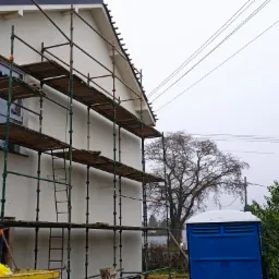 Dom jednorodzinny w trakcie prac elewacyjnych, z widocznymi rusztowaniami, świeżo położonym tynkiem i przenośną toaletą na posesji.
