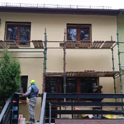 Prace na wysokościach: Dwóch pracowników na rusztowaniu przed ocieploną ścianą budynku z widocznymi oknami, jeden w neonowej czapce, drugi w ciemnym ubraniu, z częściowo pomalowaną elewacją...