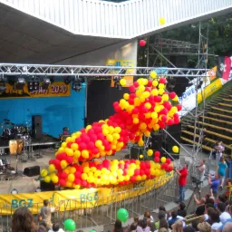 Scena plenerowa z dekoracją z czerwono-żółtych balonów, widok z góry na publiczność i fotografa dokumentującego wydarzenie. W tle instrumenty muzyczne i oświetlenie sceniczne.