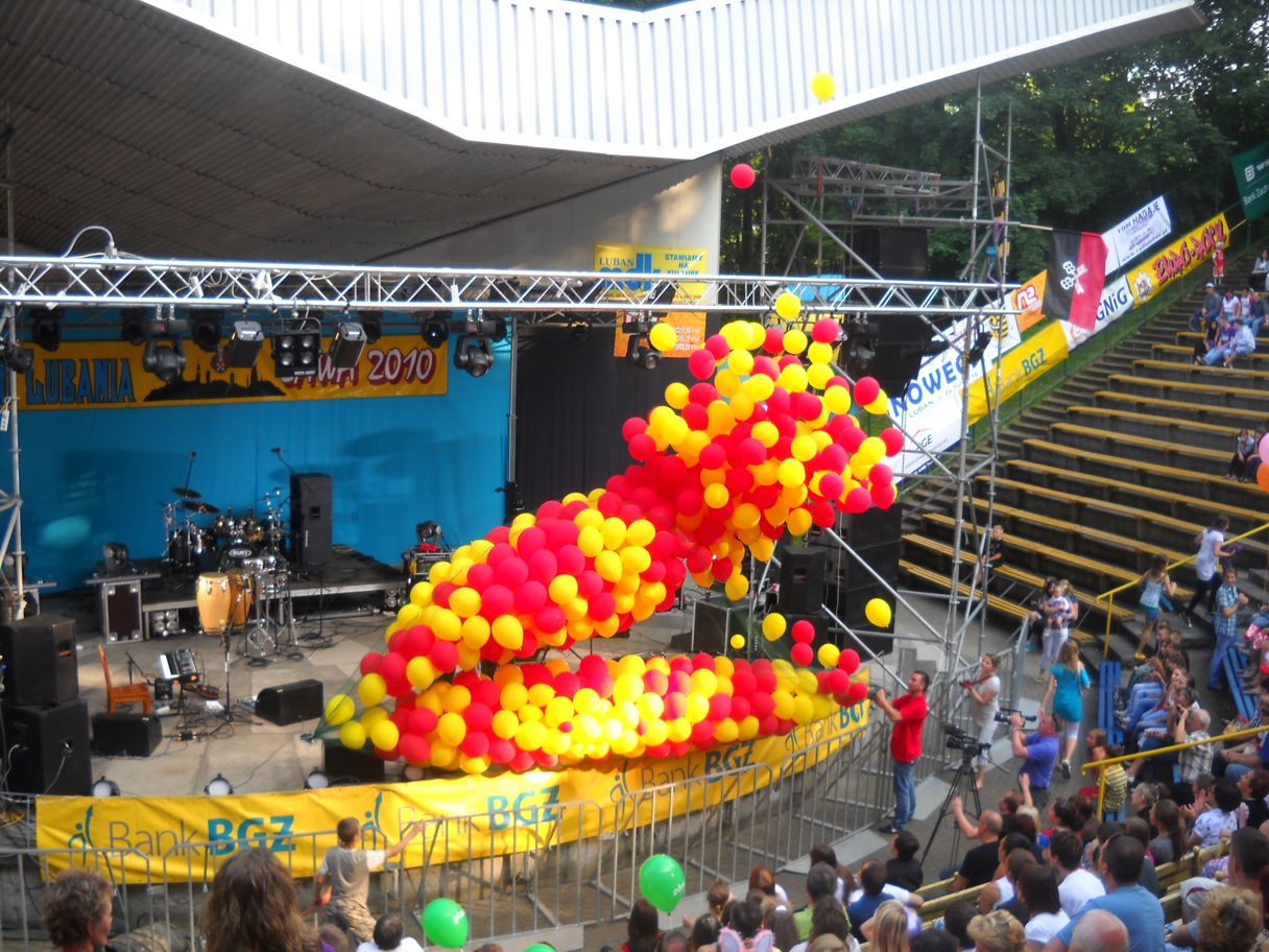 Scena plenerowa z dekoracją z czerwono-żółtych balonów, widok z góry na publiczność i fotografa dokumentującego wydarzenie. W tle instrumenty muzyczne i oświetlenie sceniczne.