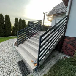 Zewnętrzne schody z jasnymi, granitowymi stopniami i ciemną, metalową balustradą, prowadzące do domu z ceglaną elewacją, widok na kamienną ścieżkę i wysokie tuje w tle.