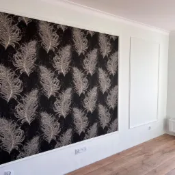 Ściana z tapetą w pióra, obramowana białą listwą, sąsiadująca z białą ścianą z dekoracyjną ramą, wykończona listwami przypodłogowymi i drewnianą podłogą.