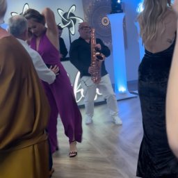 Wodzirej MIRON - Saksofonista w akcji podczas imprezy tanecznej, oświetlenie sceniczne, parkiet, ludzie w ruchu, dynamiczne ujęcie z bliska, fioletowy kombinezon.