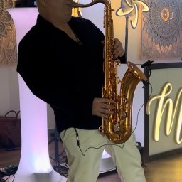 Wodzirej MIRON - Saksofonista w czarnej koszuli i białych spodniach gra na złotym saksofonie na tle dekoracji świetlnych i drewnianej ściany.
