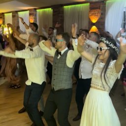 Wodzirej MIRON - Roztańczona para młoda w białych okularach i goście weselni pod błyszczącą kulą dyskotekową podczas zabawy na parkiecie. W tle ceglane ściany i okna.