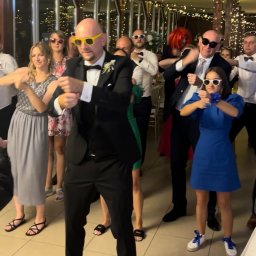 Wodzirej MIRON - Rozbawieni goście weselni w okularach tańczą w rytm muzyki. W tle dekoracje świetlne i okna. Ujęcie z perspektywy parkietu.